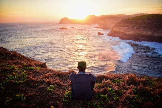 A man sitting on the ground and admiring the view of the sea and sundown