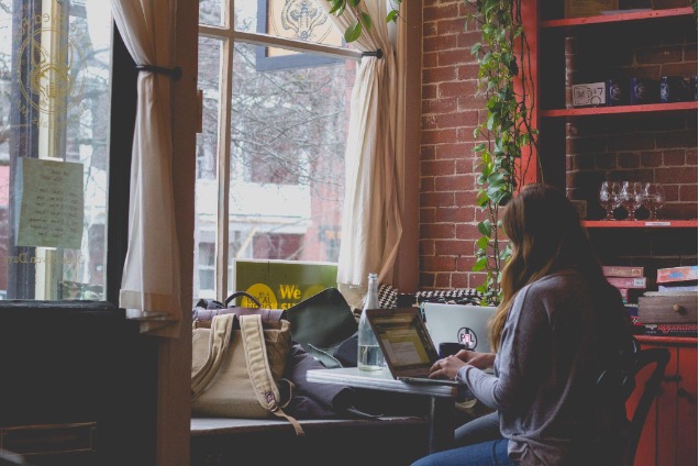 A girl sitting in a cafe next to the window and typing on a laptop.
