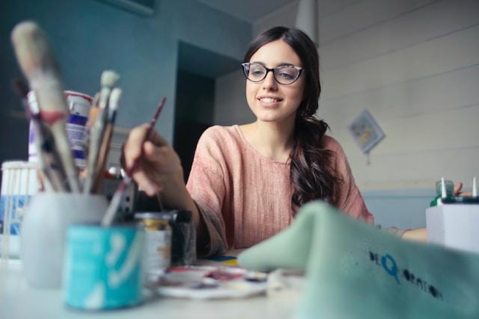 A woman with eyeglasses, in a painting studio, holding a paintbrush.